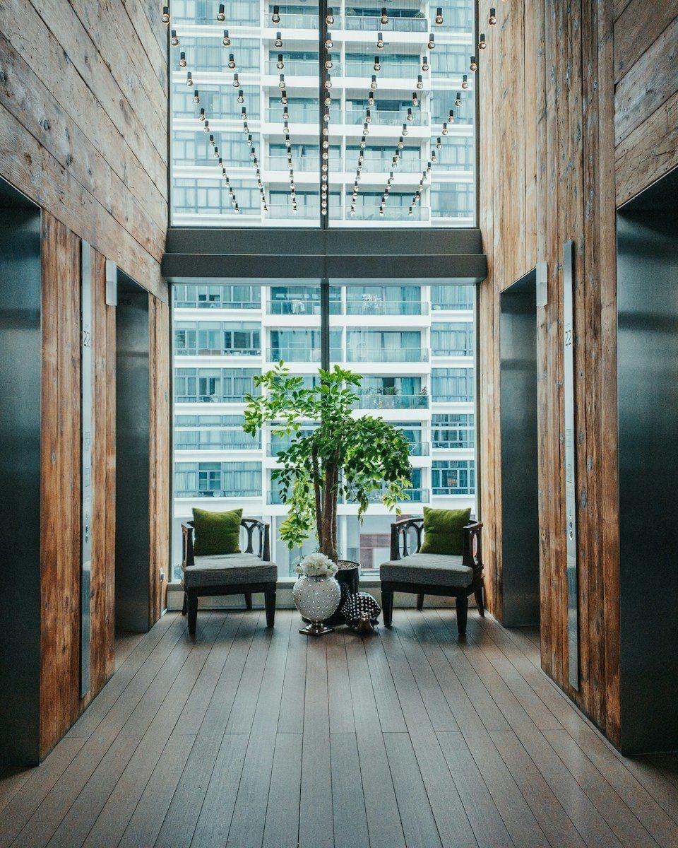 A warm elevator lobby in full color, with wood paneling, two armchairs, and a leafy tree by a tall window