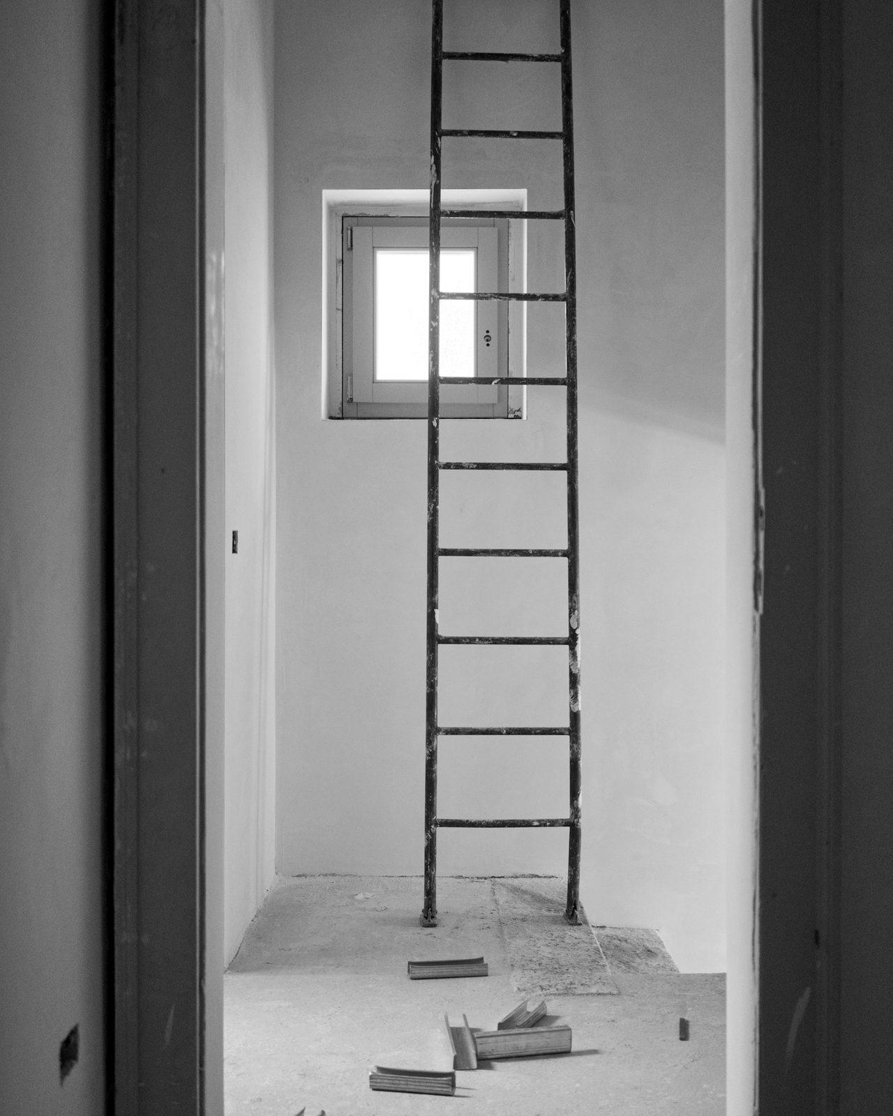 A worn metal ladder standing in a bare unfinished room, photographed in black and white